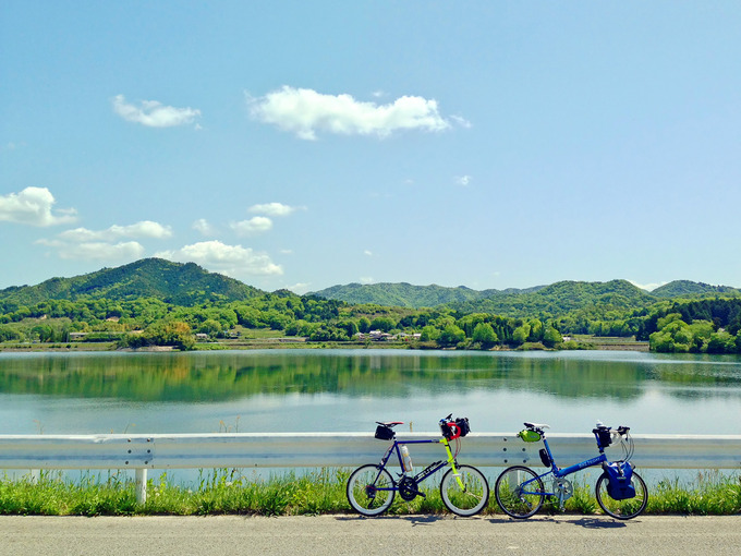 三田の青野ダムのダム湖「千丈寺湖」の風景。むこうには緑色の山々があり、湖面には山と青空が映っている。手前には2台の自転車、ニューワールドツーリストとコメットRが停められている。