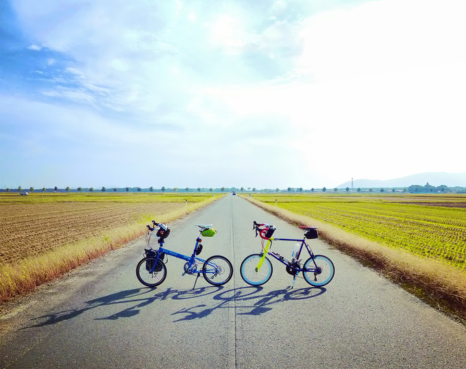 滋賀県高島市の広大な田園風景。収穫を終えた後の田んぼの真ん中に、まっすぐの道路が続いている。道路には2台の自転車、「ニューワールドツーリスト」と「コメットR」が停められている。
