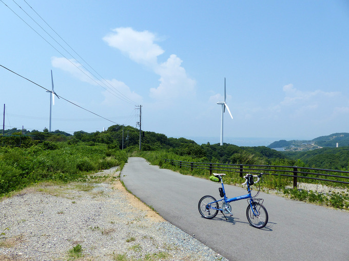 山の稜線に建てられた風力発電用の巨大な白い風車。風車が並ぶ風景をバックに、青い自転車「ニューワールドツーリスト」が停められている。稜線の合間に見える景色のはるかむこうには、青い海の水平線が少しだけ見える。