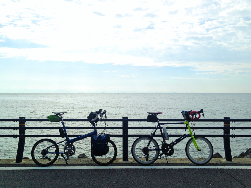 ガードレールの前に2台の自転車、バイクフライデーの折り畳みミニベロ「ニューワールドツーリスト」とミニベロロード「コメットR」が停められている。そのむこうには海が広がっている。