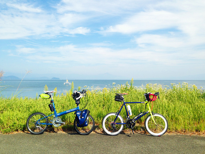 滋賀県高島市新旭の「木津」交差点付近から見る琵琶湖の風景。緑色の草むらのむこうに、青い琵琶湖の水面が広がっている。手前にはバイクフライデーの折り畳みミニベロ「ニューワールドツーリスト」とフジ「コメットR」が停められている。