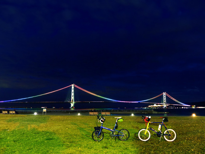 日没後の明石の大蔵海岸公園で見る、明石海峡大橋の写真。電飾が点灯して七色に光っている。手前の広場には2台の自転車、バイクフライデーの折り畳みミニベロ「ニューワールドツーリスト」とミニベロロード「コメットR」が停められている。