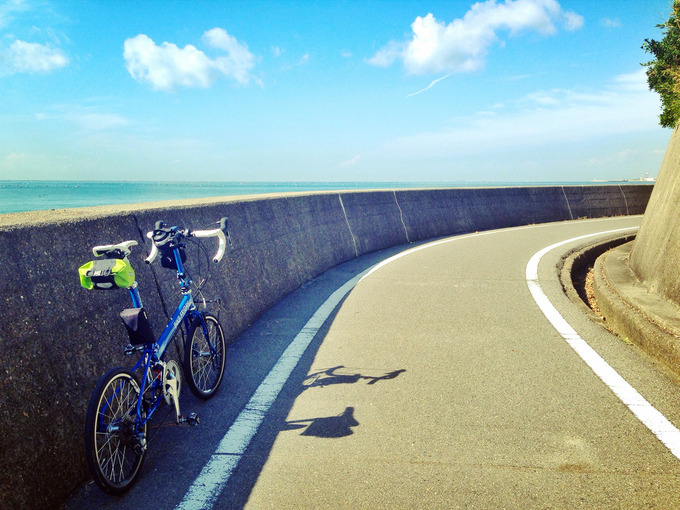 兵庫県明石市内の「播磨サイクリングロード」の写真。右にカーブした道の左側には青い海が広がっている。カーブの外側の壁の横に、バイクフライデーの折り畳みミニベロ「ニューワールドツーリスト」が停められている。