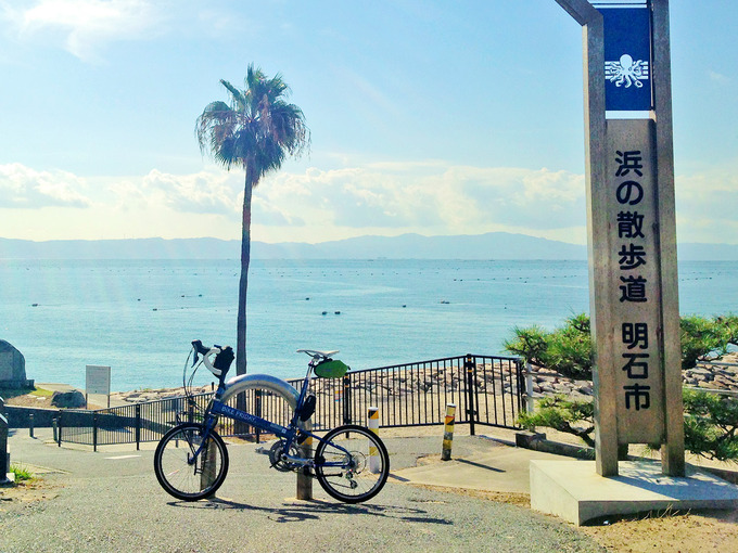 播磨サイクリングロードの江井ヶ島付近の写真。少し高台になっていて、「浜の散歩道・明石市」と書かれた看板がある。むこうには海と椰子の木が見える。手前にはバイクフライデーの折り畳みミニベロ「ニューワールドツーリスト」が停められている。