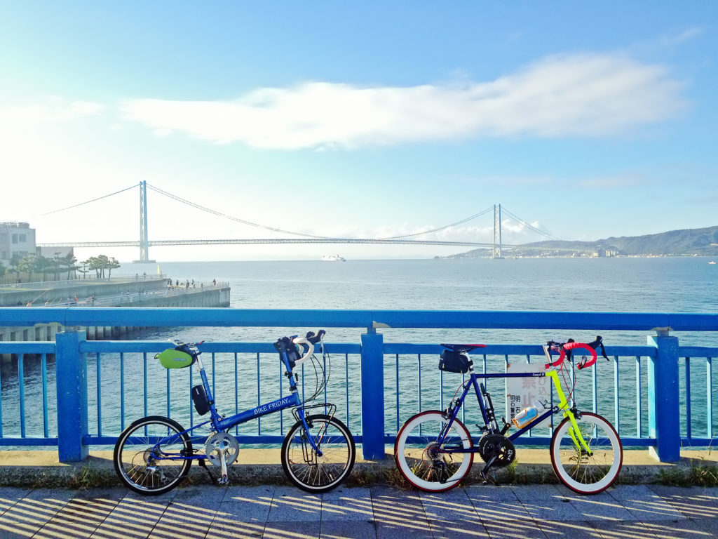 大蔵海岸付近の、朝霧川にかかる橋の上に2台の自転車が停められている写真。背景には海と淡路島、明石海峡大橋が見える。