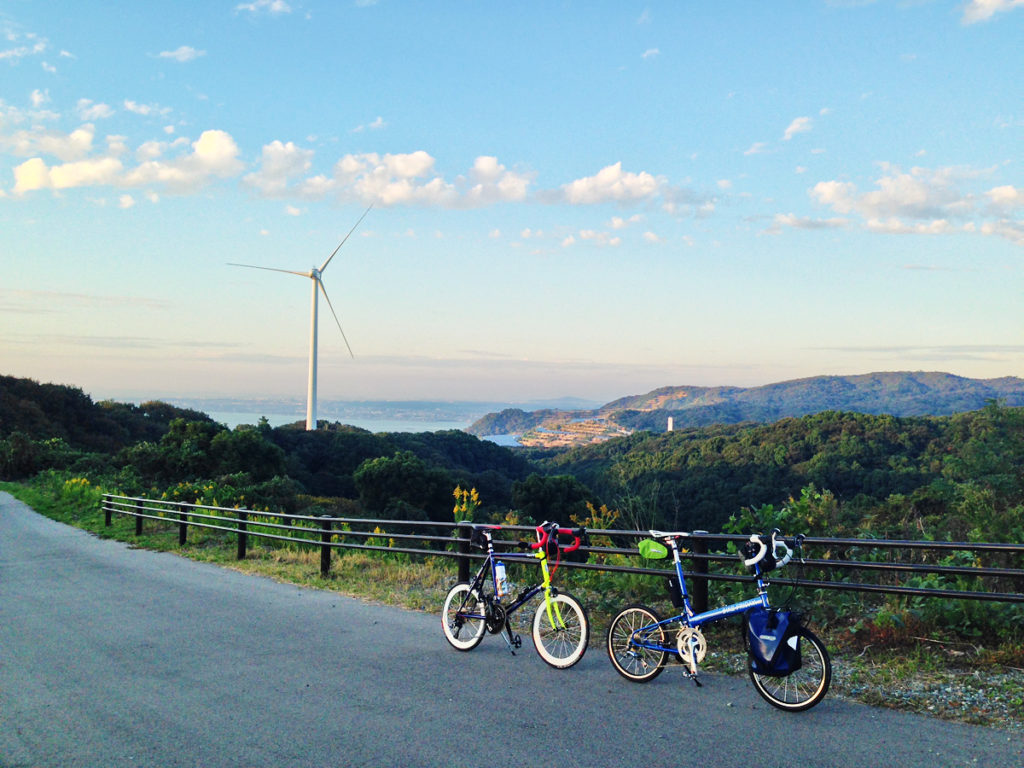 淡路島の山上の風景に2台のミニベロが停められている写真。後ろに発電風車が見える。