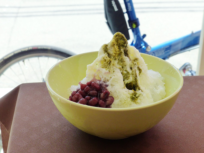 薄緑色の深い器に、かき氷が入っている写真。緑色の抹茶や白いミルクがかけられて、赤茶色の小豆が添えられている。