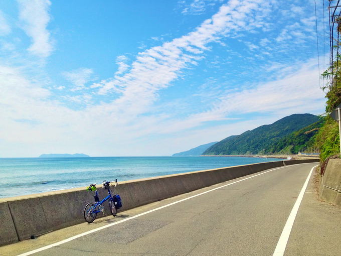 淡路島の南端付近の海岸線の風景。海岸線の道路「南淡路水仙ライン」から見る青い海、青い空、秋の雲の風景で、道路にはバイクフライデーの折り畳みミニベロ「ニューワールドツーリスト」が停められていて、海の向こうには小さな離島「沼島」が見える。