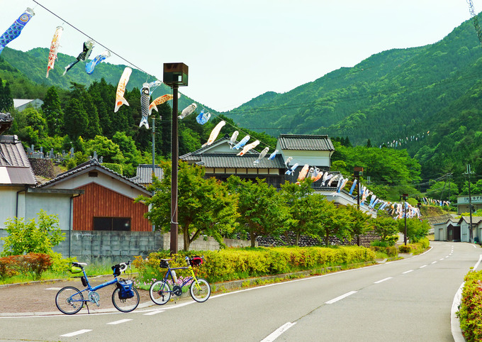 兵庫県神崎郡神河町にある「グリーンエコー笠形」に続く道路に沿って、たくさんの「鯉のぼり」が道しるべとして吊るされている風景。ずっと先まで鯉のぼりの列が続いている。道路には2台の自転車、「ニューワールドツーリスト」と「コメットR」が停められている。