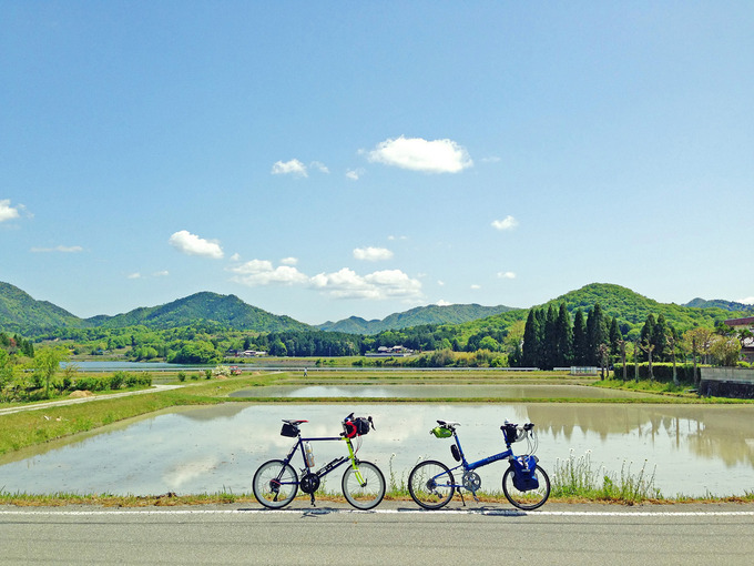 三田の田園風景の写真。田植え前の水が張られた田んぼに空や雲が映っている。むこうには緑色の山々が見える。手前には2台の自転車、ニューワールドツーリストとコメットRが停められている。