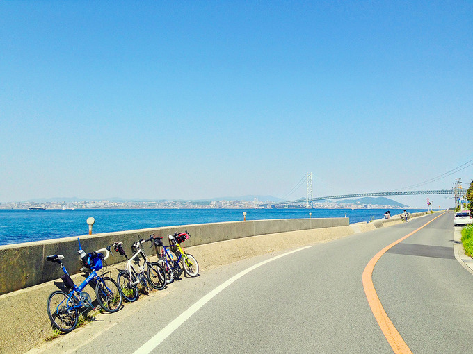 淡路島・松帆の浦付近の風景の写真。左側には青い海が広がり、前方には明石海峡大橋が見える。道路脇には3台のミニベロが停められている。